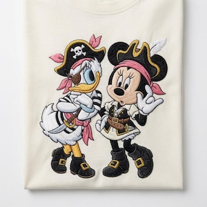 May include: A cream-coloured t-shirt featuring a large, detailed embroidery of two cartoon characters dressed as pirates. One character wears a black pirate hat with a skull and crossbones, an eyepatch, and a striped shirt. The other wears a black pirate hat with a white feather and a black and white outfit.