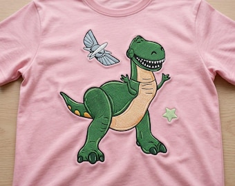May include: A light pink short-sleeve t-shirt featuring a large embroidered graphic of a green dinosaur with a tan belly. Above the dinosaur is a small white and blue winged emblem, and to the right is a small, light green embroidered star.