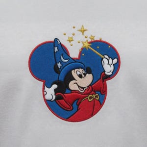 Op de afbeelding: Een geborduurde patch op een witte stoffen achtergrond met Mickey Mouse als tovenaar. Het personage draagt een blauwe hoed met witte sterren en een maansikkel, en een rode mantel. Mickey houdt een gouden toverstaf met gele sterren omhoog.