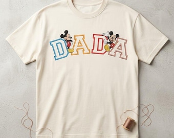 May include: A cream-colored short-sleeve t-shirt featuring the word DADA in large, colorful block letters. The letters are outlined in blue, yellow, red, and brown. Mickey Mouse figures are positioned between the letters on the shirt.