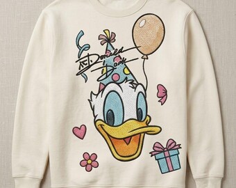 May include: A cream-colored crewneck sweatshirt featuring an embroidered graphic of Donald Duck wearing a polka-dot party hat. The design includes a tan balloon, a blue gift box with a pink bow, a pink flower, a heart, and the text Donald Duck.