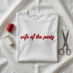 Puede incluir: Camiseta blanca de cuello redondo con el texto bordado "wife of the party" en rojo. La camiseta está colocada sobre una superficie de tela blanca, con carretes de hilo rojo y blanco, agujas y tijeras cerca.