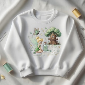 Könnte beinhalten: Weißes Sweatshirt mit einer farbenfrohen Illustration einer Fee, eines Schmetterlings und eines Baumhauses. Die Fee trägt ein grünes Kleid und Flügel. Eine Flasche mit Trank und Sterne sind ebenfalls abgebildet. Das Sweatshirt ist aus weichem Material.