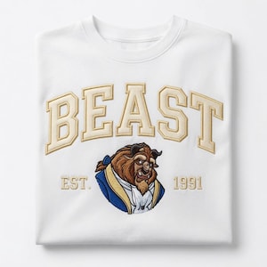 May include: A white crewneck sweatshirt featuring an embroidered design. The word BEAST is stitched in large, beige, block-style letters. Below, an embroidered character portrait of a beast in a blue coat, with the text EST. 1991 in beige.