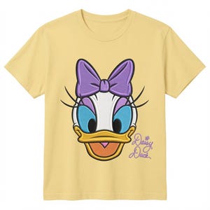 May include: A pale yellow short-sleeve t-shirt featuring a large embroidered graphic of Daisy Duck's face. The character has a purple bow, blue eyelids, and a yellow beak. The text Daisy Duck is embroidered in purple script on the lower right.