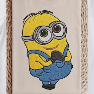 Peut inclure: Un t-shirt beige avec une broderie détaillée à la machine représentant un personnage Minion jaune. Le personnage porte une salopette bleue, des gants noirs et des lunettes à monture argentée avec un œil vert et un œil marron, en position debout.
