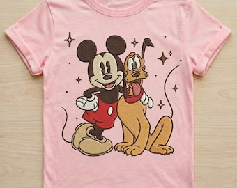 May include: A light pink short-sleeve t-shirt featuring a central embroidered graphic of Mickey Mouse and Pluto. Mickey is wearing red shorts and white gloves, while Pluto has a red collar. The design includes small brown star accents.