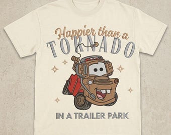 May include: A cream-colored t-shirt featuring a graphic of a brown tow truck character with a smiling face and buck teeth. The text reads "Happier than a TORNADO IN A TRAILER PARK" in brown and gray lettering, accented by small four-pointed star icons.