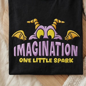 May include: A black t-shirt featuring an embroidered graphic of a purple dragon with yellow horns and wings peeking out from behind the word IMAGINATION in large purple letters, with the text ONE LITTLE SPARK in yellow underneath.