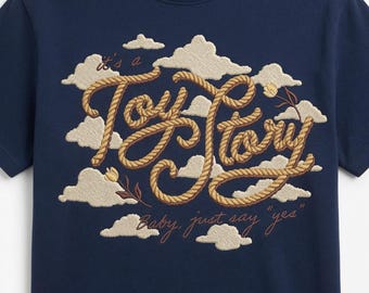 May include: A navy blue short-sleeved t-shirt featuring an embroidered design of beige clouds and the text "it's a Toy Story" rendered in a rope-like font, with small floral accents and the additional text "Baby, just say "yes"" below.