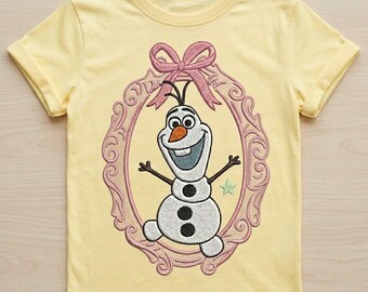 May include: A pale yellow short-sleeved t-shirt featuring an embroidered design of a white snowman with an orange carrot nose and three black buttons. The snowman is centered inside an oval pink frame topped with a pink bow and a small star.