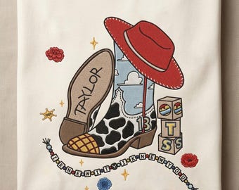 May include: An off-white t-shirt featuring an embroidered design of a cow-print cowboy boot, a red hat, and blocks with the letters T, S, and S. The text "TAYLOR" is on the boot sole, and a beaded string reads "I KNEW IT I KNEW YOU".