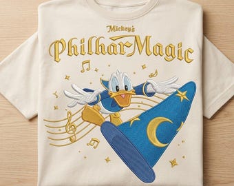 May include: A cream-colored t-shirt featuring an embroidered graphic of Donald Duck wearing a blue wizard hat with a yellow crescent moon and stars. The text reads Mickey's PhilharMagic in gold, accompanied by gold musical notes and staff lines.