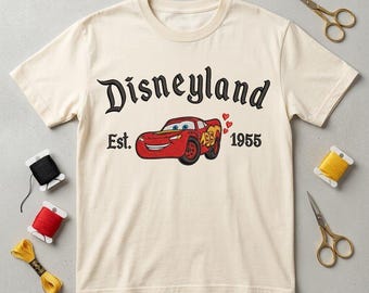 May include: A cream-colored short-sleeve t-shirt featuring an embroidered red race car graphic, three small red hearts, and the black text Disneyland Est. 1955. The design is centered on the chest of the shirt, which is displayed flat.