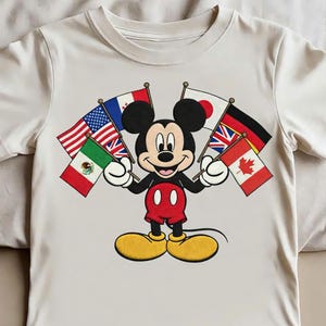 May include: A cream-colored short-sleeve t-shirt featuring a graphic of Mickey Mouse holding several international flags, including those of the United States, Mexico, France, Japan, the United Kingdom, Germany, and Canada, in his white-gloved hands.