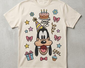 May include: A cream-colored short-sleeve t-shirt featuring a graphic of Goofy wearing a party hat. The design includes a birthday cake with three candles, pink bows, colorful stars, a gift box, a cupcake, and the handwritten text Goofy.