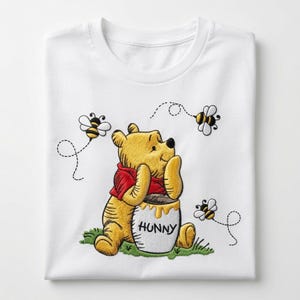 May include: A white t-shirt featuring an embroidered graphic of a yellow bear wearing a red shirt, sitting on green grass. The bear is leaning on a white pot labeled HUNNY, with three small black and yellow bees flying around it.