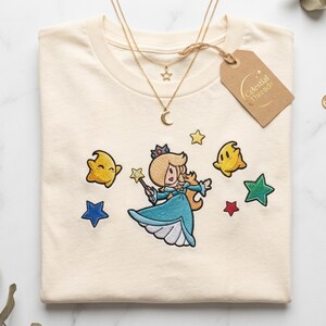Könnte beinhalten: Ein cremefarbenes T-Shirt mit einer Stickerei eines blonden Charakters in einem blauen Kleid, der einen Sternenstab hält, umgeben von gelben, blauen, roten und grünen Sternen. Eine goldfarbene mehrlagige Halskette und ein braunes Etikett Celestial Threads.