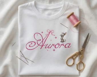 May include: A white t-shirt featuring pink embroidered cursive text that reads Aurora, accented with a small pink bow, four-pointed stars, and a small brown rabbit illustration. A spool of pink thread and metal scissors are placed nearby.