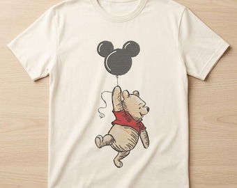 May include: A cream-colored short-sleeve t-shirt featuring a central graphic of a light brown bear wearing a red shirt, holding a string attached to a black balloon shaped like a mouse head. The design uses a sketch-like illustration style.
