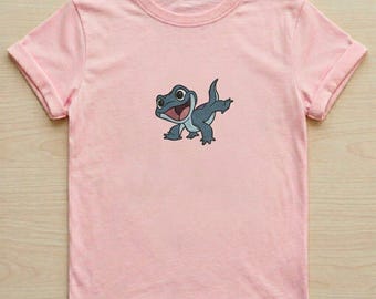 May include: A light pink short-sleeved t-shirt featuring a centered graphic of a small, dark blue lizard with large eyes and a wide, open-mouthed smile. The lizard is depicted in a playful pose with one leg raised, showcasing a light blue underbelly.