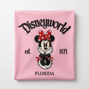 May include: A light pink t-shirt featuring black embroidered text that reads Disneyworld, est. 1971, and FLORIDA. Centered is an embroidered graphic of Minnie Mouse wearing a red and white polka dot bow, black ears, and white gloves, resting her chin on her hands.
