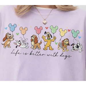 Könnte beinhalten: Ein hellviolettes Sweatshirt mit einer Grafik von sechs Zeichentrickhunden, die jeweils einen verschiedenfarbigen, Mickey-Maus-förmigen Luftballon halten. Darunter steht der Text "Life is better with dogs" in schwarzer Schreibschrift.