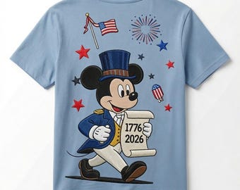 May include: A light blue t-shirt featuring an embroidered graphic of Mickey Mouse wearing a top hat and colonial-style coat. Mickey holds a scroll with the text 1776 2026. The design includes a small American flag, fireworks, and red and blue stars.