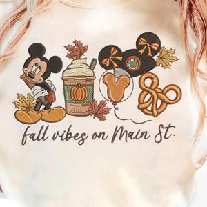 May include: A cream-colored t-shirt featuring an embroidered design of Mickey Mouse, a pumpkin-topped coffee cup, mouse-ear headbands, a balloon, and a pretzel, surrounded by autumn leaves. The text reads, fall vibes on Main St.