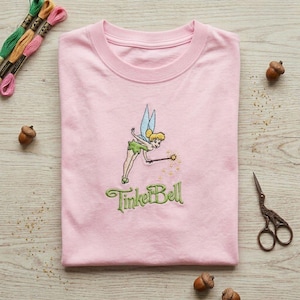 Könnte beinhalten: Hellrosa Rundhals-T-Shirt mit einem gestickten Bild von Tinker Bell mit einem Zauberstab und dem Text "Tinker Bell" in Grün. Stickgarn und Eicheln sind sichtbar.