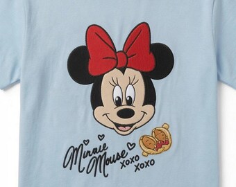 May include: A light blue t-shirt featuring an embroidered graphic of Minnie Mouse with a red bow. Below the character, the text reads Minnie Mouse XOXO, accompanied by small heart icons and a small illustration of two tan-colored food items.
