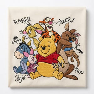 Könnte beinhalten: Ein cremefarbener Stoff mit einer Stickerei der Winnie Puuh-Figuren, darunter Tigger, Rabbit, I-Aah, Ferkel, Kanga und Roo. Die Namen der einzelnen Figuren sind mit schwarzem Garn neben den jeweiligen Charakteren aufgestickt.