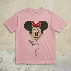 May include: A light pink short-sleeve t-shirt featuring an embroidered graphic of Minnie Mouse as a balloon with a red and white polka dot bow. The text Minnie Mouse is embroidered in black script below the balloon, and a tag is at the neck.