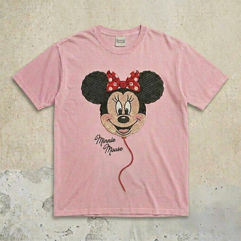 May include: A light pink short-sleeve t-shirt featuring an embroidered graphic of Minnie Mouse as a balloon with a red and white polka dot bow. The text Minnie Mouse is embroidered in black script below the balloon, and a tag is at the neck.