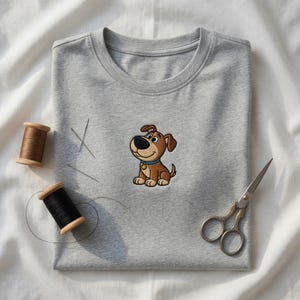 Könnte beinhalten: Ein graues T-Shirt mit einer Stickerei eines Cartoon-Hundes. Der Hund ist braun mit einem blauen Halsband und einem goldenen Anhänger. Nähutensilien, darunter Garnrollen, Nadeln und eine Schere, sind um das T-Shirt auf einem weißen Stoffhintergrund angeordnet.