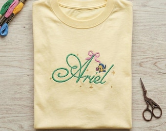 May include: A light yellow t-shirt with the name "Ariel" embroidered in green cursive, adorned with a pink bow and a small cartoon fish. Embroidery floss and scissors are visible.