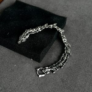 May include: A silver-colored dragon bracelet with a detailed design. The bracelet features a dragon head at one end and a clasp at the other. The dragon's body is articulated, and the bracelet is resting on a black surface.