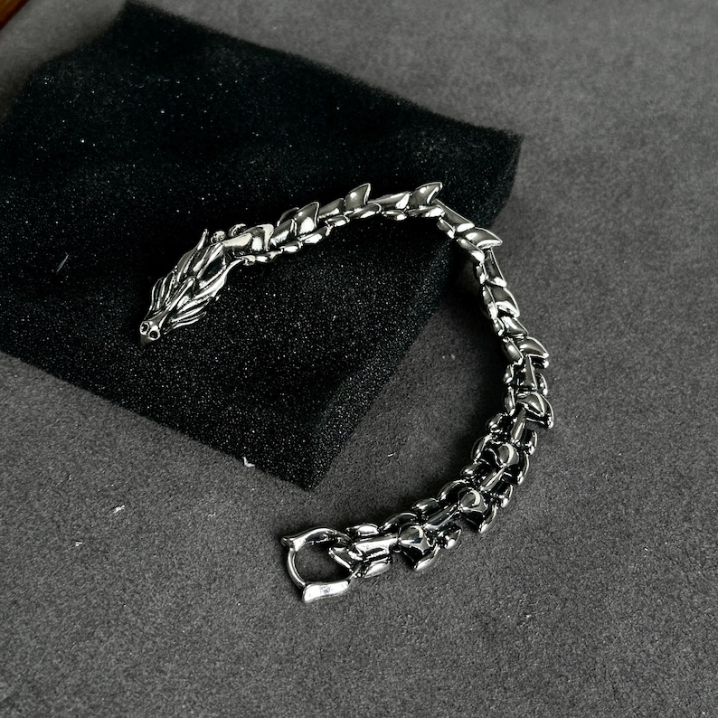May include: A silver-colored dragon bracelet with a detailed design. The bracelet features a dragon head at one end and a clasp at the other. The dragon's body is articulated, and the bracelet is resting on a black surface.