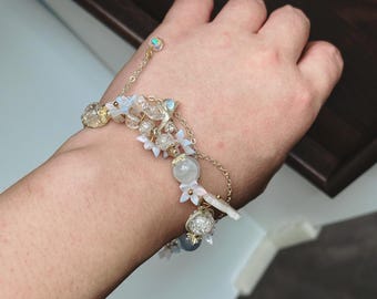 Puede incluir: Una pulsera de cadena dorada de múltiples hilos con cuentas de vidrio craquelado transparente, esferas translúcidas lisas y pequeños dijes en forma de flor blanca. Incluye una cadena ajustable delicada con una pequeña cuenta iridiscente.