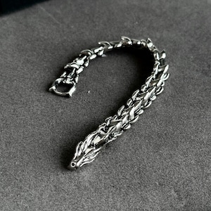May include: A silver-colored bracelet with a dragon-scale design. The bracelet features a detailed dragon head at one end and a clasp at the other. The links are intricately designed, giving the bracelet a textured appearance. The bracelet is displayed on a gray surface.