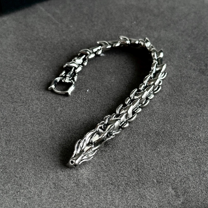 May include: A silver-colored bracelet with a dragon-scale design. The bracelet features a detailed dragon head at one end and a clasp at the other. The links are intricately designed, giving the bracelet a textured appearance. The bracelet is displayed on a gray surface.