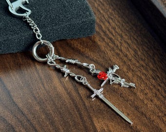 Puede incluir: Un llavero de metal plateado con un cierre de mosquetón, una cadena y dos dijes colgantes. Un dije es una espada larga y delgada, y el otro es una cruz adornada con una pequeña rosa roja brillante en el centro. Ambos cuelgan de alambre de espino.