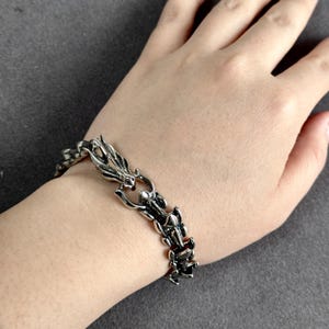 May include: Silver-toned metal bracelet featuring a dragon design. The bracelet is composed of interconnected dragon-shaped links, creating a flexible and detailed accessory. The dragon's head and tail are visible, adding to the intricate design.