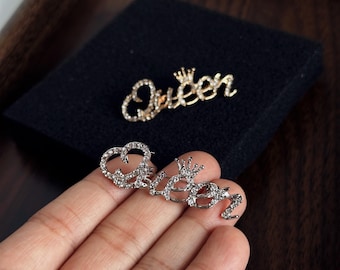 Puede incluir: Dos broches de metal dorado con la palabra Queen en letra cursiva y un pequeño icono de corona sobre la letra n. Cada broche está incrustado con brillantes transparentes que cubren toda la superficie de las letras.