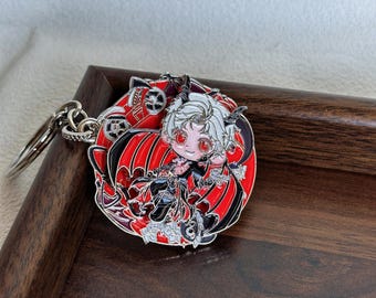 Puede incluir: Un llavero de metal circular que presenta un personaje estilizado con cabello blanco, ojos rojos y cuernos pequeños. El personaje viste un atuendo negro y rojo con alas, sobre un fondo rojo vibrante con detalles intrincados de engranajes.