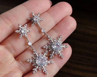Puede incluir: Un par de pendientes colgantes de tono plateado con diseño de copo de nieve. Cada pendiente consta de dos dijes en forma de copo de nieve conectados por una pequeña piedra redonda transparente. Los copos tienen pequeños cristales claros.