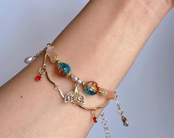Puede incluir: Una pulsera de dijes en tono dorado con un dije central de cola de sirena, dos cuentas de vidrio craquelado en azul y naranja, pequeñas perlas blancas y diminutos cristales rojos. La pulsera incluye una cadena delicada y un cierre de mosquetón.