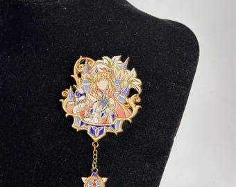 Puede incluir: Un broche de metal esmaltado en tono dorado que presenta un personaje estilizado con cabello largo, vistiendo un atuendo blanco y azul, rodeado de elementos florales. Una pequeña cadena dorada cuelga de la parte inferior, unida a un dije triangular con detalles en azul y rojo.