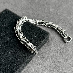 May include: A silver-colored dragon bracelet with intricate detailing. The bracelet features a dragon head at one end and a clasp at the other. The dragon's body is made up of interconnected segments, creating a flexible design. The bracelet is displayed on a black surface.