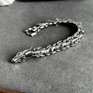 May include: A silver-colored dragon bracelet. The bracelet is made of metal and features a dragon head at one end and a clasp at the other. The dragon's body is segmented, and the bracelet is lying on a gray surface.